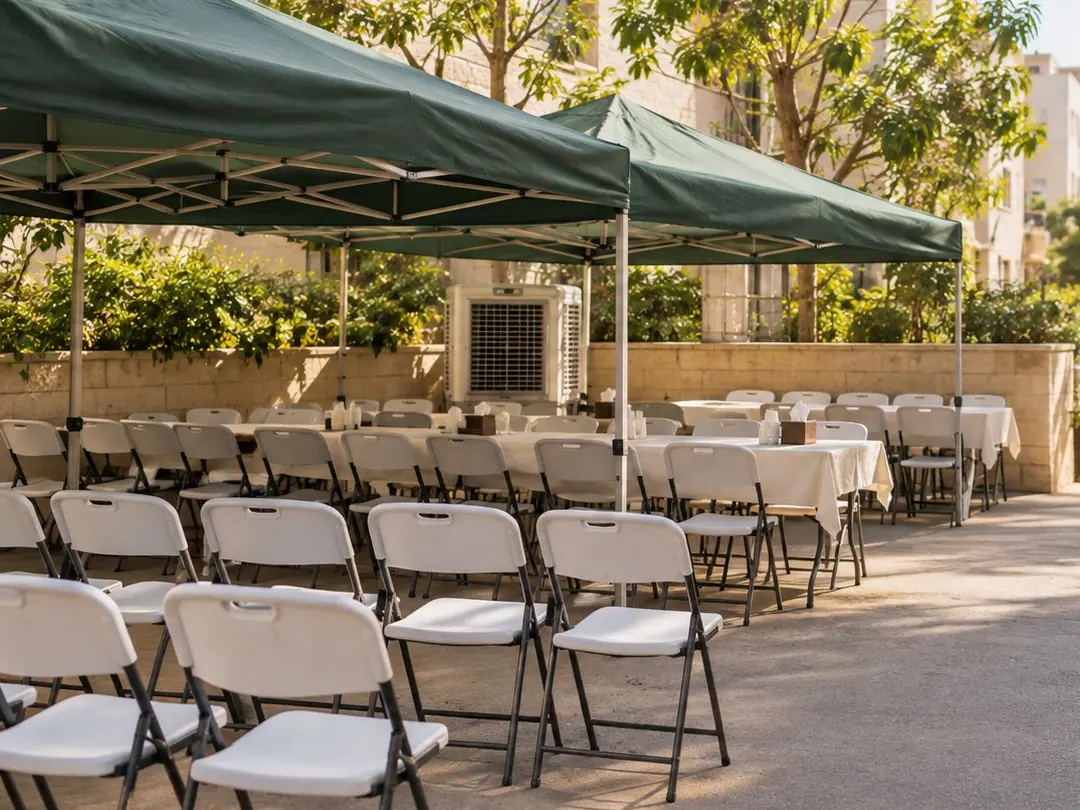צליות ירוקות בחצר בין בנייני אבן, ומתחתן שולחנות עם מפות לבנות, שורות כיסאות מתקפלים ומצנן